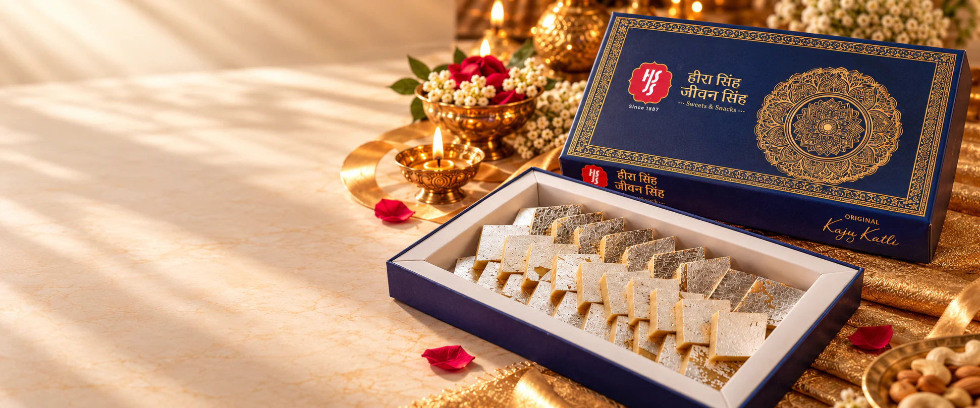 A navy-and-gold HSJS Original Kaju Katli box, open to show silver-leaf kaju katli, with brass diyas and flowers behind it