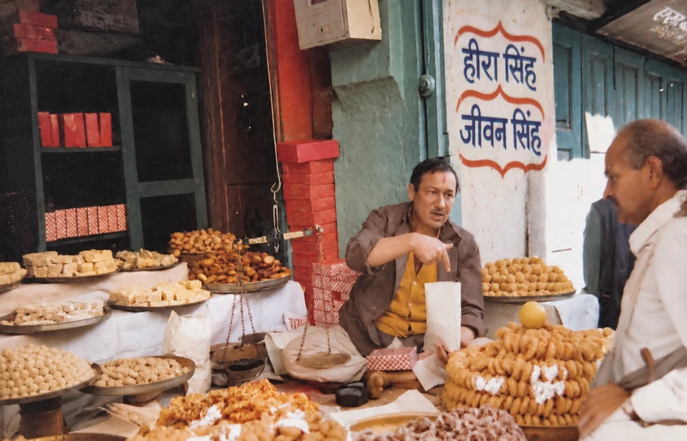 Jeewan Singh seated cross-legged at the counter among trays of sweets, gesturing as he talks to a customer, the shop name painted on the wall behind him