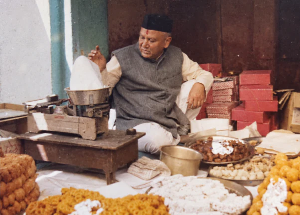 Archive photograph of an elder in a black cap seated cross-legged behind trays of mithai, red sweet boxes stacked behind him