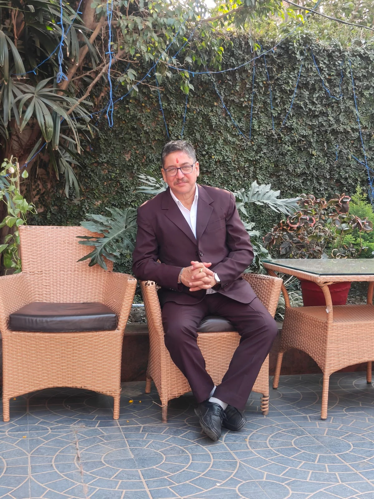 Navneet Singh in a dark maroon suit and glasses, seated in a wicker chair in a garden