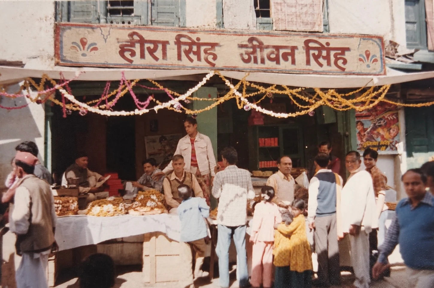 The shopfront under its painted Heera Singh Jeewan Singh signboard, strung with garlands, with sweets laid out on a table and customers gathered